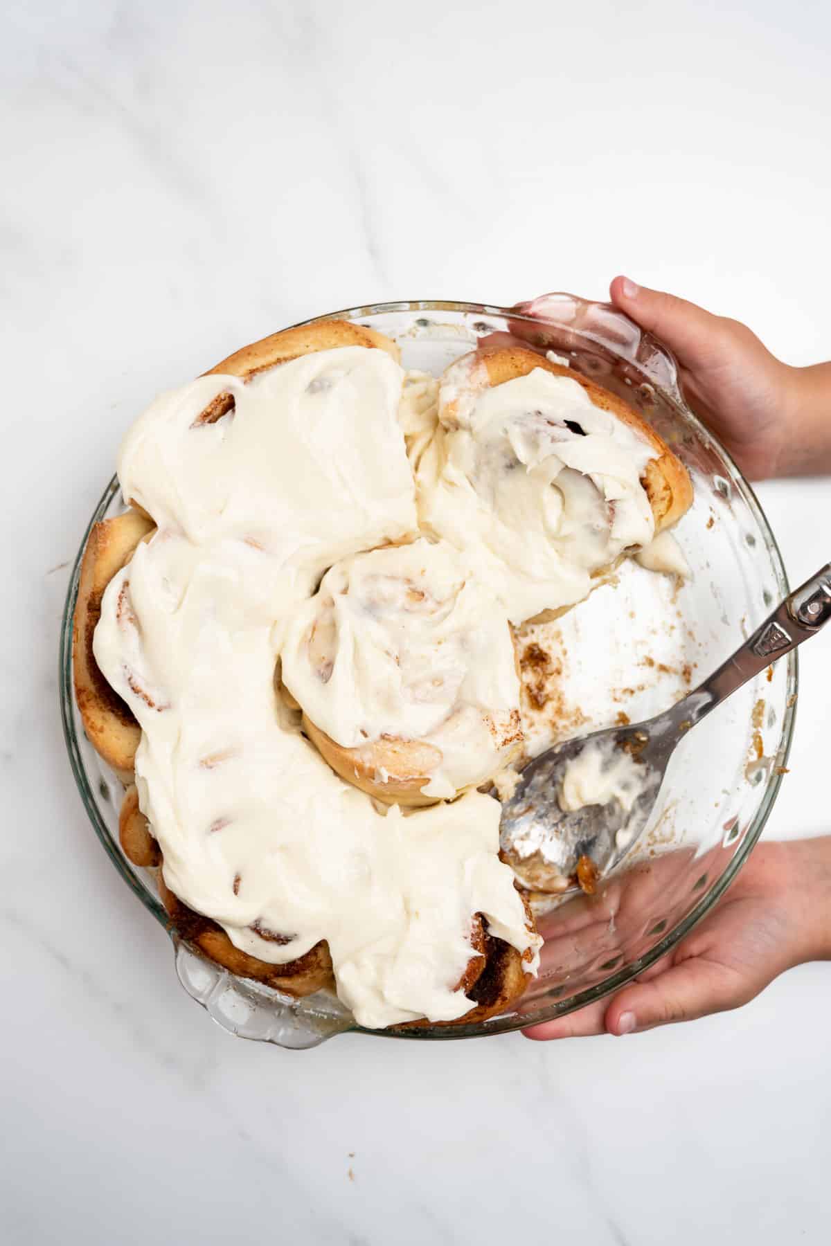 two hands holding homemade cinnamon rolls in a round baking pan with spoon.