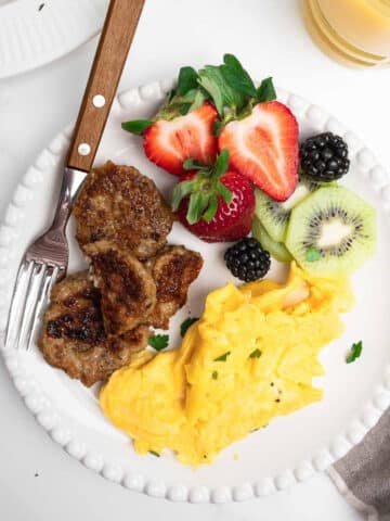 chicken sausage, fresh fruit and scrambled eggs on a white plate with a fork.
