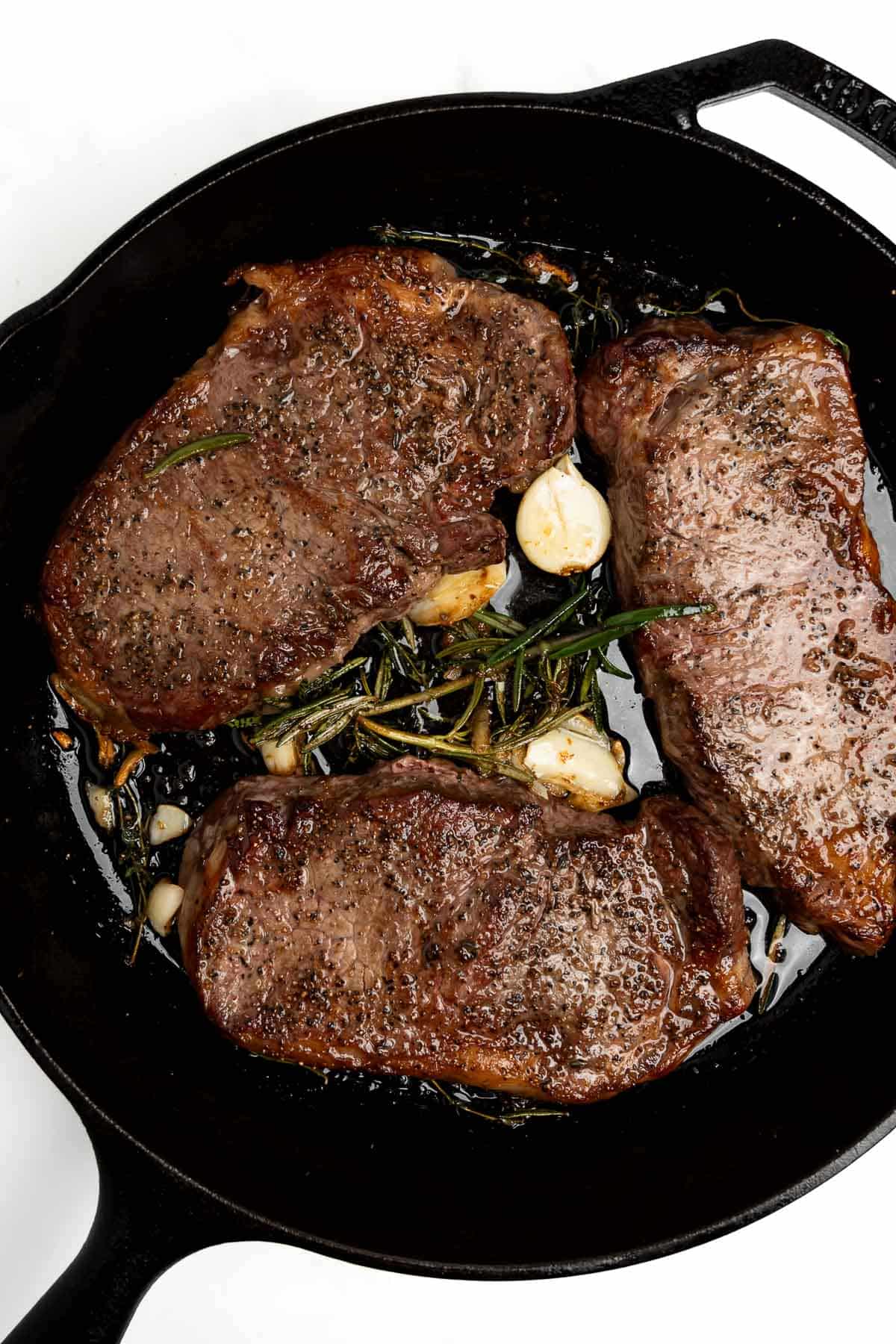 three steaks in a cast iron skillet with garlic and herbs.