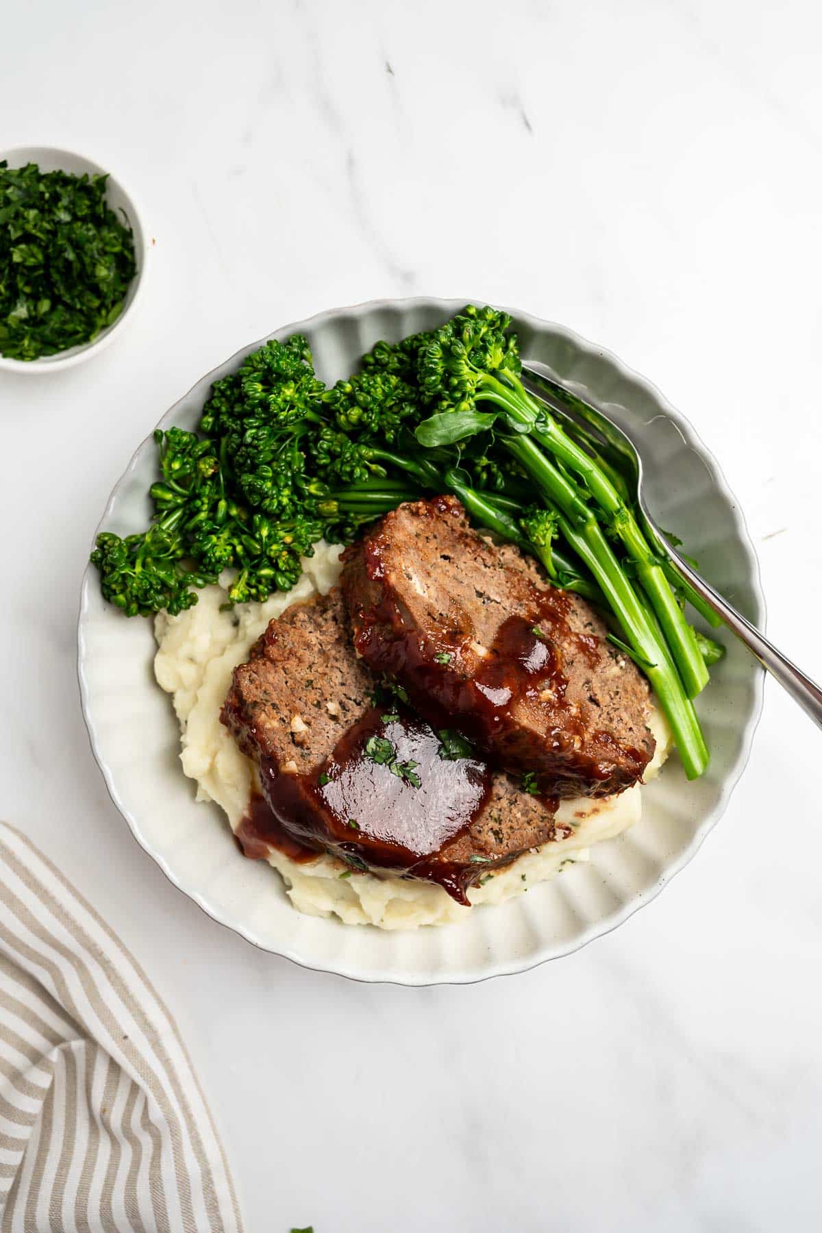 sliced meatloaf on a plate over mashed potatoes with baby broccoli