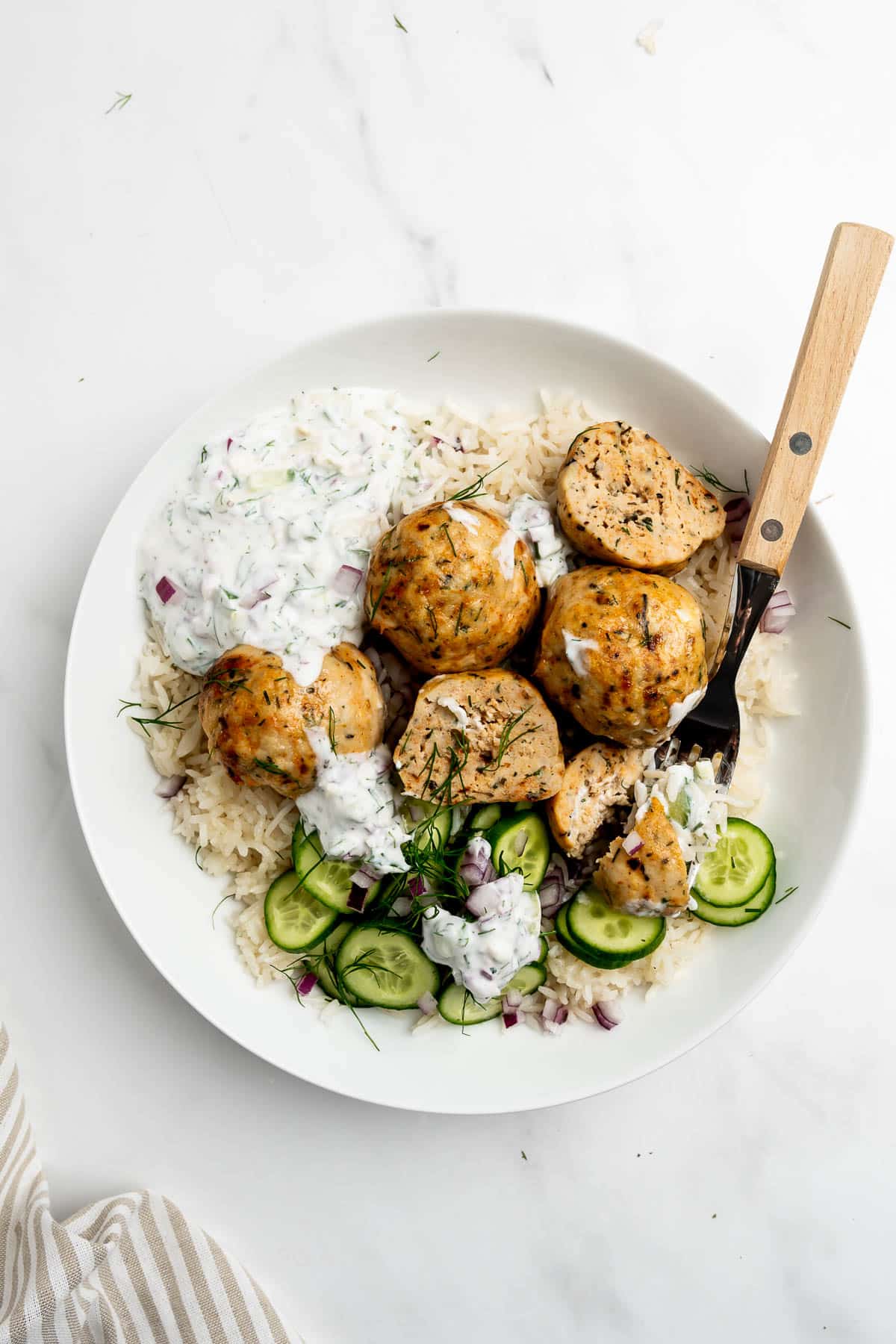Greek chicken meatballs in a bowl with rice, sliced cucumber, and tzatziki sauce.